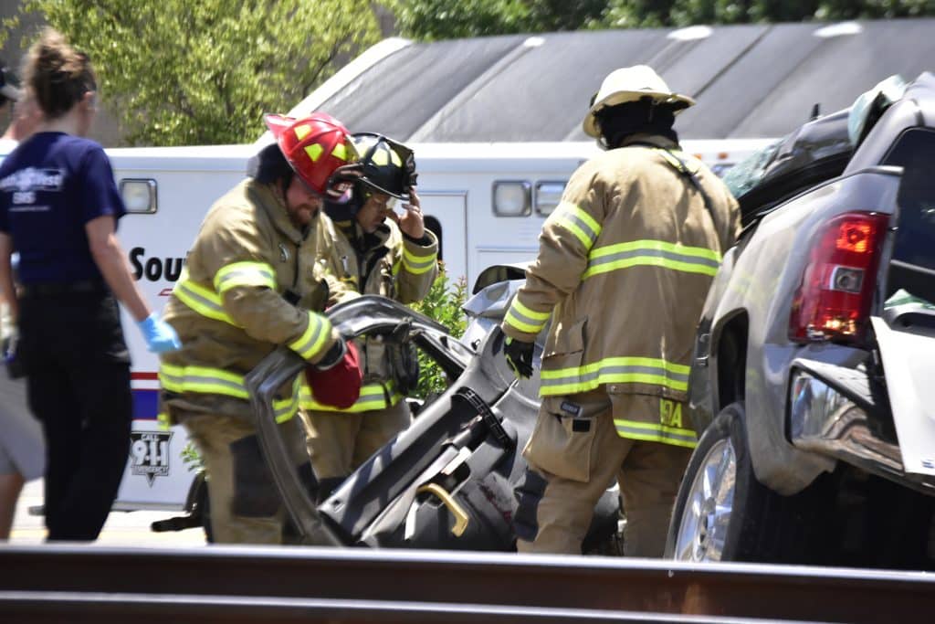 Firefighters in turnout gear work at a vehicle crash inspecting a damaged car with rescue tools nearby an ambulance is visible in the background