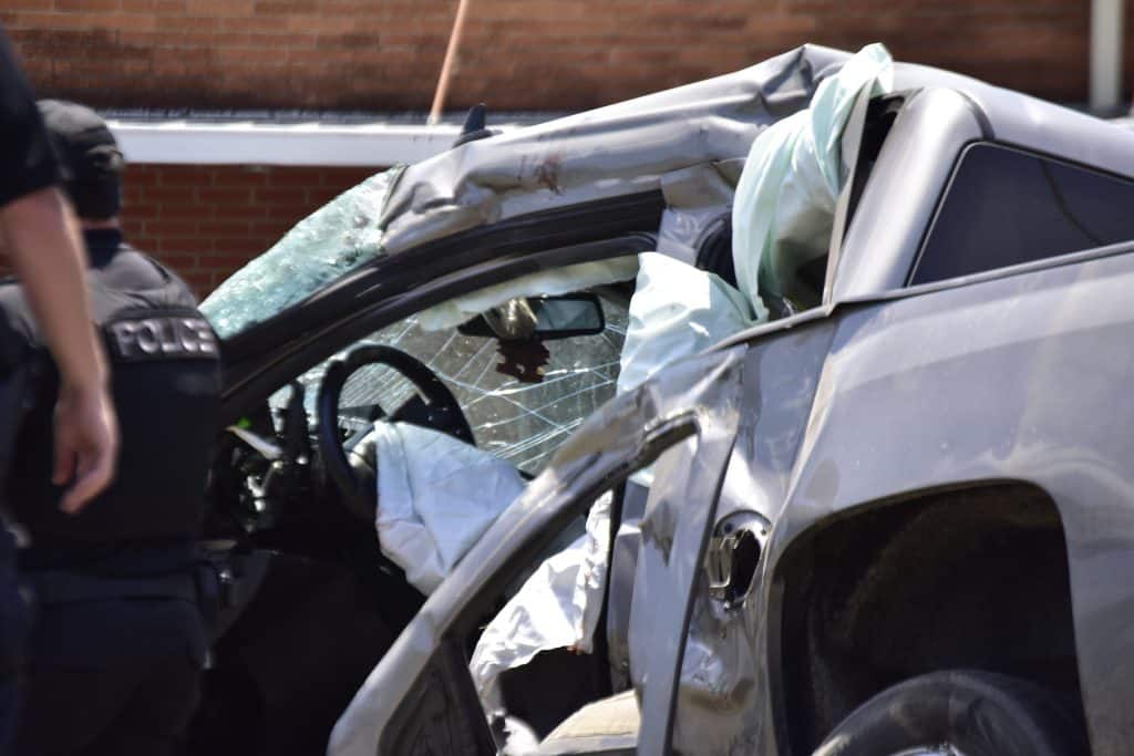 Police officer stands beside a heavily damaged silver SUV from a crash with a cracked windshield and deployed airbags visible on the passenger side