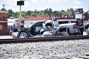 Two heavily damaged vehicles crash near a storefront, with bystanders and a police officer inspecting the scene.